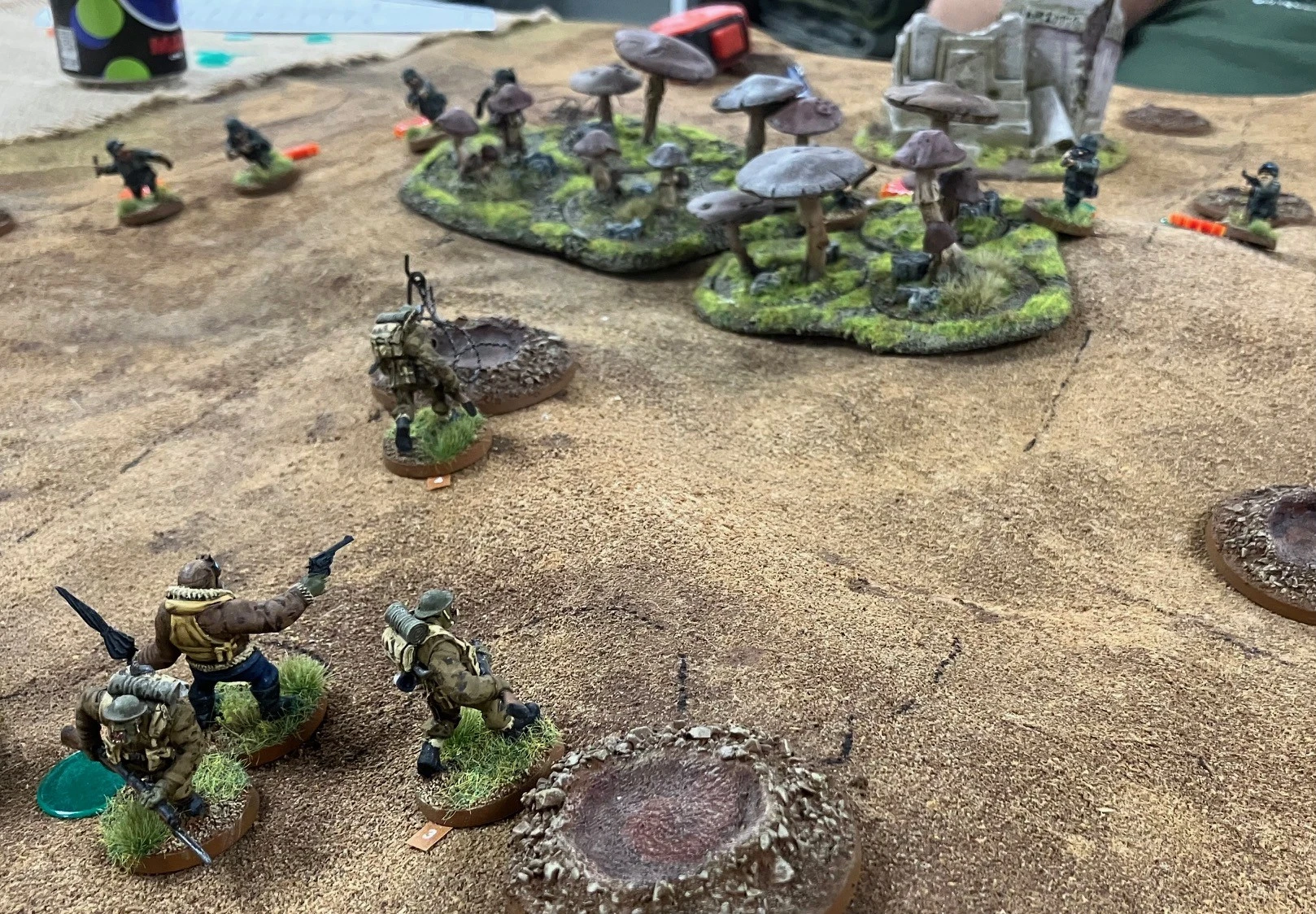 A tabletop gaming scene features several miniature figures positioned on a textured, sandy terrain. In the foreground, two figures are visible: one is a muscular character holding a pistol, while the other carries a backpack and appears to be in a tactical stance. Behind them, a cluster of large, stylized mushrooms and a rocky outcrop create a vibrant backdrop. The overall setting suggests a fantasy or sci-fi theme, with elements of nature and rugged terrain enhancing the atmosphere.