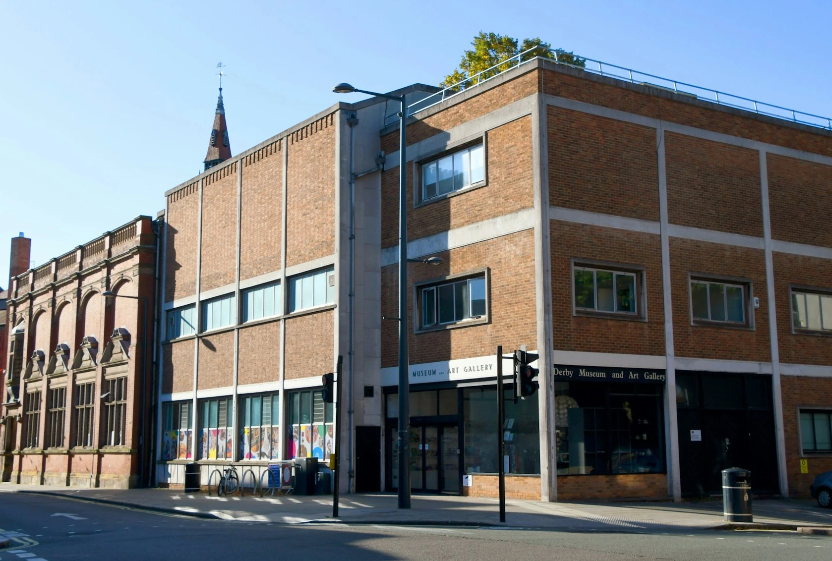 The image shows the exterior of the Derby Museum and Art Gallery, a modern brick building with large windows. The entrance features a sign that reads "Derby Museum and Art Gallery," and there are bicycles parked nearby. In the background, a church spire is visible, rising above the museum, indicating the urban setting of the location. The scene is well-lit, suggesting a clear day.