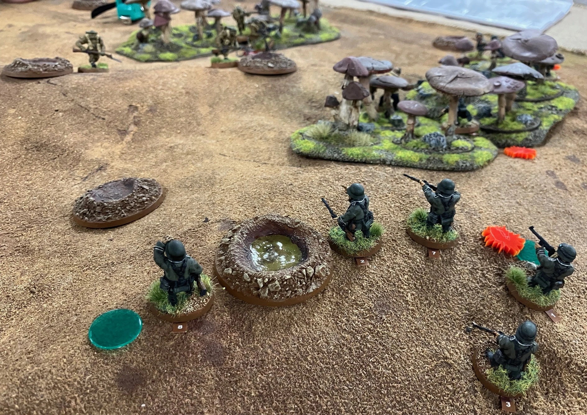 A tabletop gaming scene featuring several miniature soldiers positioned on a textured, sandy terrain. In the foreground, two soldiers in green uniforms stand near a circular, rocky depression filled with murky water, while another soldier is seen in the background, moving towards a cluster of mushrooms. The environment includes various terrain features, such as grassy patches and additional miniature figures scattered throughout the scene.