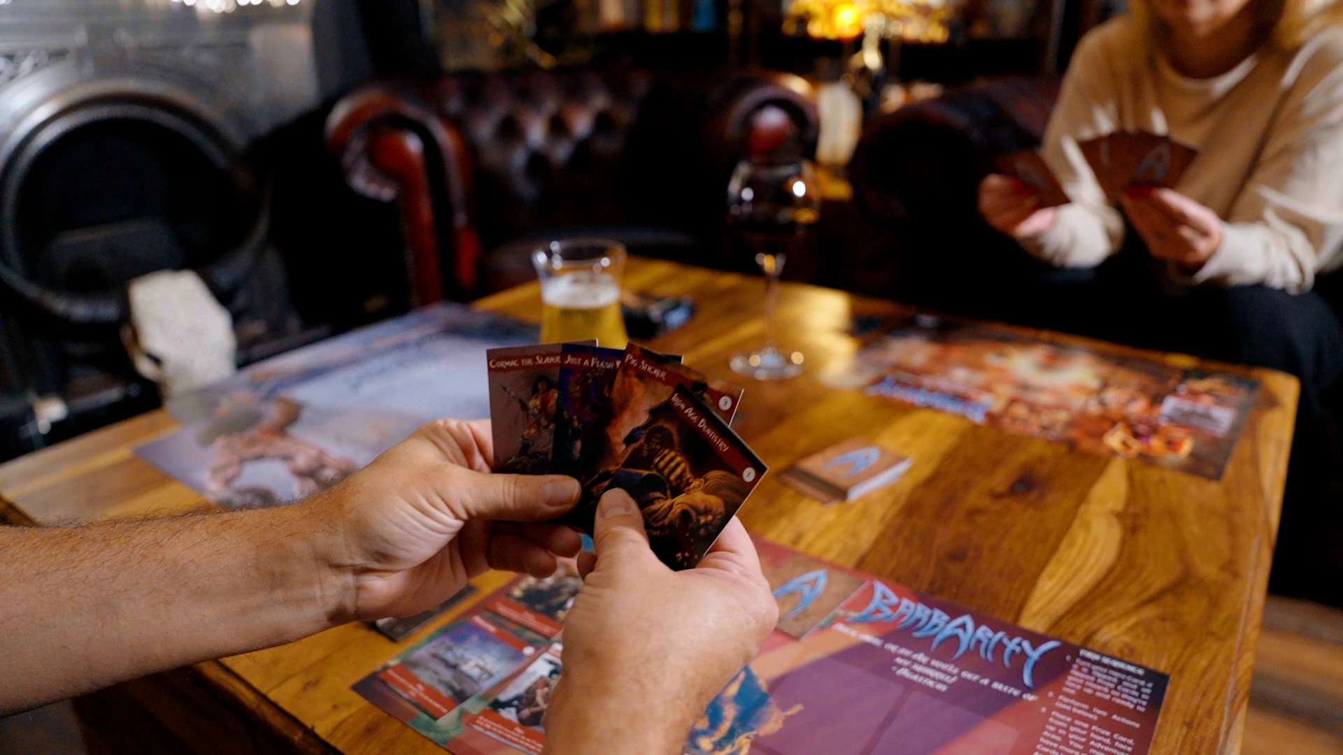 The image features a pair of hands holding three colorful game cards, each depicting different characters or actions related to the game "Barbarity." The background shows a wooden table with additional game cards spread out, along with a glass of beer and a wine glass. In the blurred background, a cozy, dimly lit pub setting is visible, complete with a leather armchair and a fireplace. The overall atmosphere suggests a casual gaming session in a lively tavern.