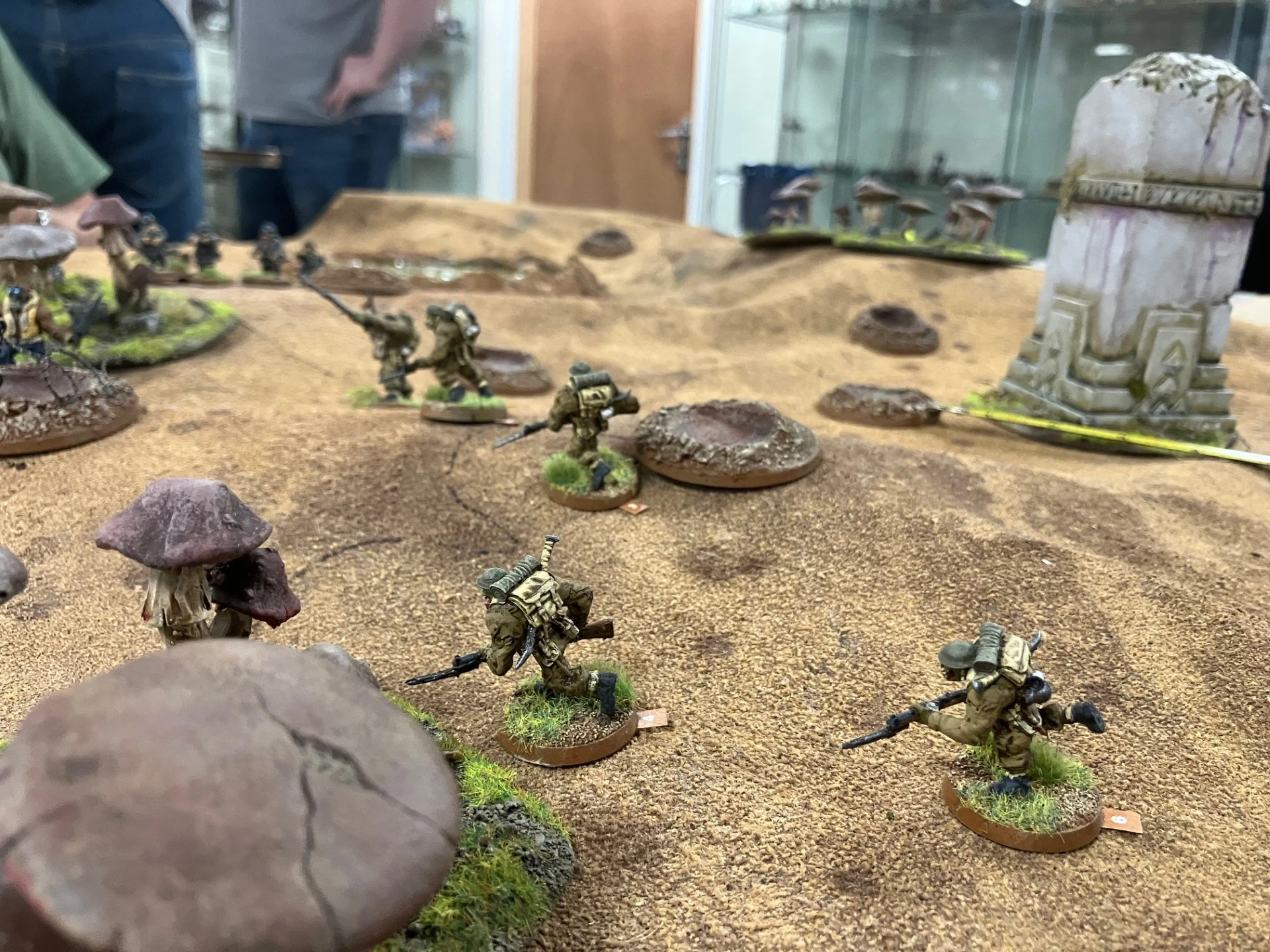 A tabletop gaming scene features several miniature figures representing soldiers in a fantasy setting, positioned on a textured, sandy terrain with patches of green grass. The soldiers are equipped with rifles and wear camouflage uniforms, while some are advancing in a line, suggesting a tactical movement. In the background, large, stylized mushrooms and a stone structure add to the whimsical atmosphere of the game.