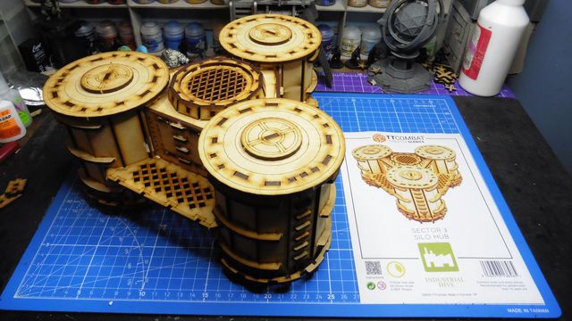 A detailed model of the Sector 3 Silo Hub from TT Combat is prominently displayed on a blue cutting mat. The structure features multiple cylindrical silos with intricate wooden textures and circular tops, showcasing a combination of open latticework and solid panels. In the background, various paint pots and a few other miniature models are visible, along with the instruction sheet for the Silo Hub, which includes an illustration of the assembled model.