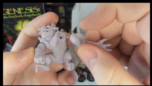 A person holds a detailed miniature of a creature from Fenris Games' Wyrdworld series, showcasing its intricate features such as muscular limbs and a fierce expression. The miniature is unpainted and made of a light-colored material, emphasizing its sculpted details. In the background, a colorful game box titled "GENESIS: The First Book of Nightmares" is partially visible, along with other miniatures displayed on a shelf.