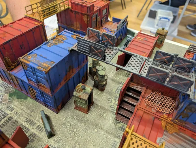 Angled overhead view of a miniature tabletop terrain model depicting an industrial or sci-fi setting. The scene includes multiple stacked shipping containers, painted in weathered blue, red, and brown tones. Some containers have open doors or missing panels, revealing interior spaces. A metal walkway with railings connects several containers at an upper level. Below, a smaller structure resembling a control box or generator sits on the ground, along with cylindrical objects resembling barrels or fuel canisters. A metal staircase with a mesh tread leads to the upper walkway. The ground surface appears textured to resemble concrete or pavement, with scattered debris. No text or people are visible.
