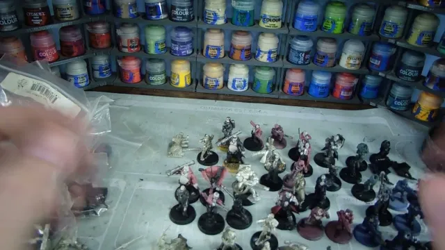 A variety of Heresy Miniatures models are arranged on a work surface, showcasing a mix of painted and unpainted figures in various poses. In the background, shelves filled with paint pots in a range of colors, including blues, greens, and reds, provide a vibrant backdrop. The models include soldiers and fantastical characters, some with detailed bases, while the surface beneath them is marked with paint splatters and tools of the hobby.