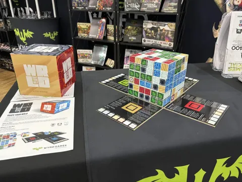 A wooden cube puzzle game on a black table with six colored cube types—forest, ocean, cave, desert, lava, tundra—partially assembled. Two game boards with numbered tiles and a red scoring token lie nearby. Shelves in background display other board game boxes.
