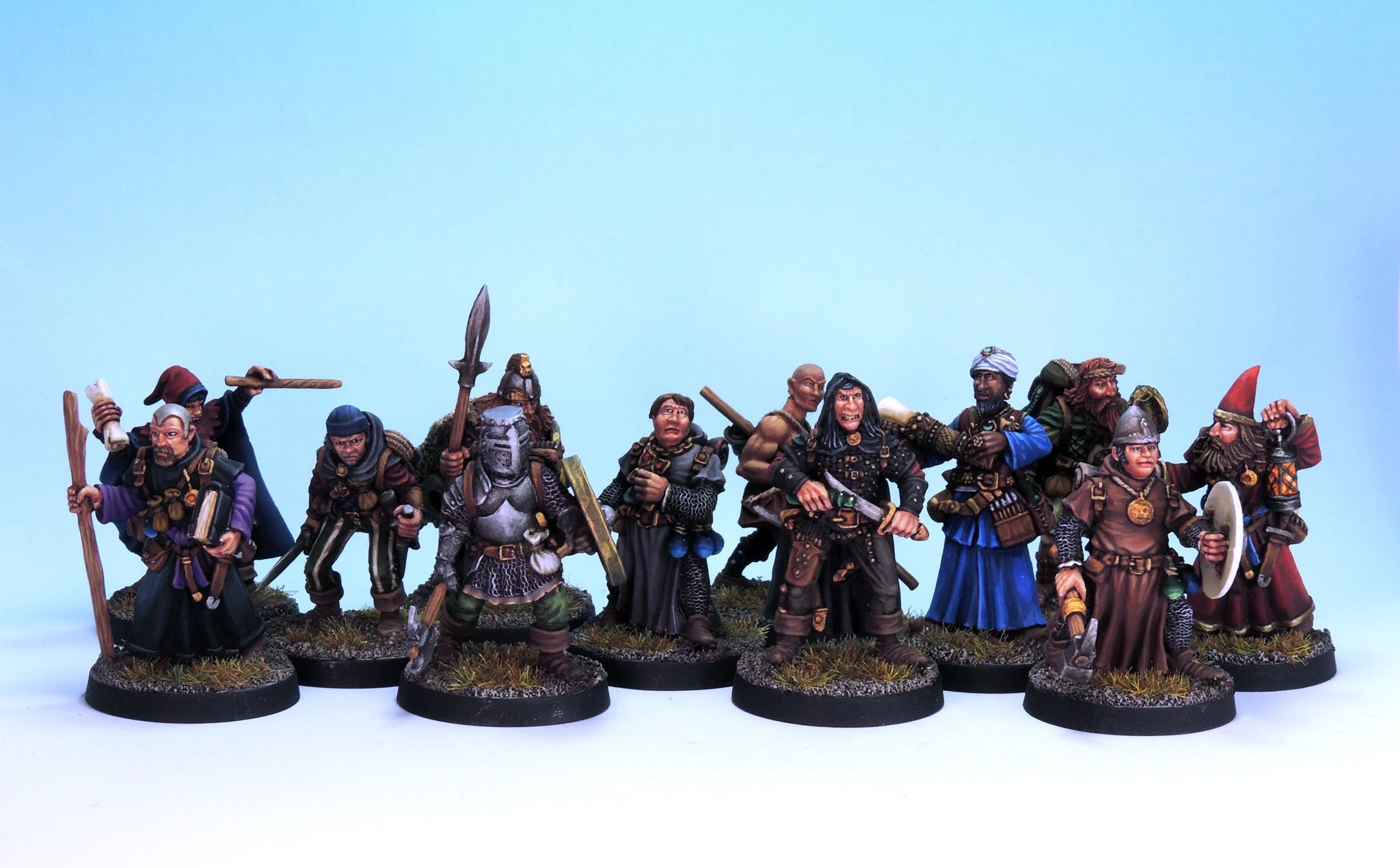 A diverse group of painted miniatures stands against a light blue background, showcasing various adventurers and characters. The figures include a mix of warriors, spellcasters, and rogues, each detailed with unique clothing, weapons, and expressions. They are mounted on circular bases with tufts of grass, highlighting their fantasy theme and adding to the overall aesthetic of the collection.