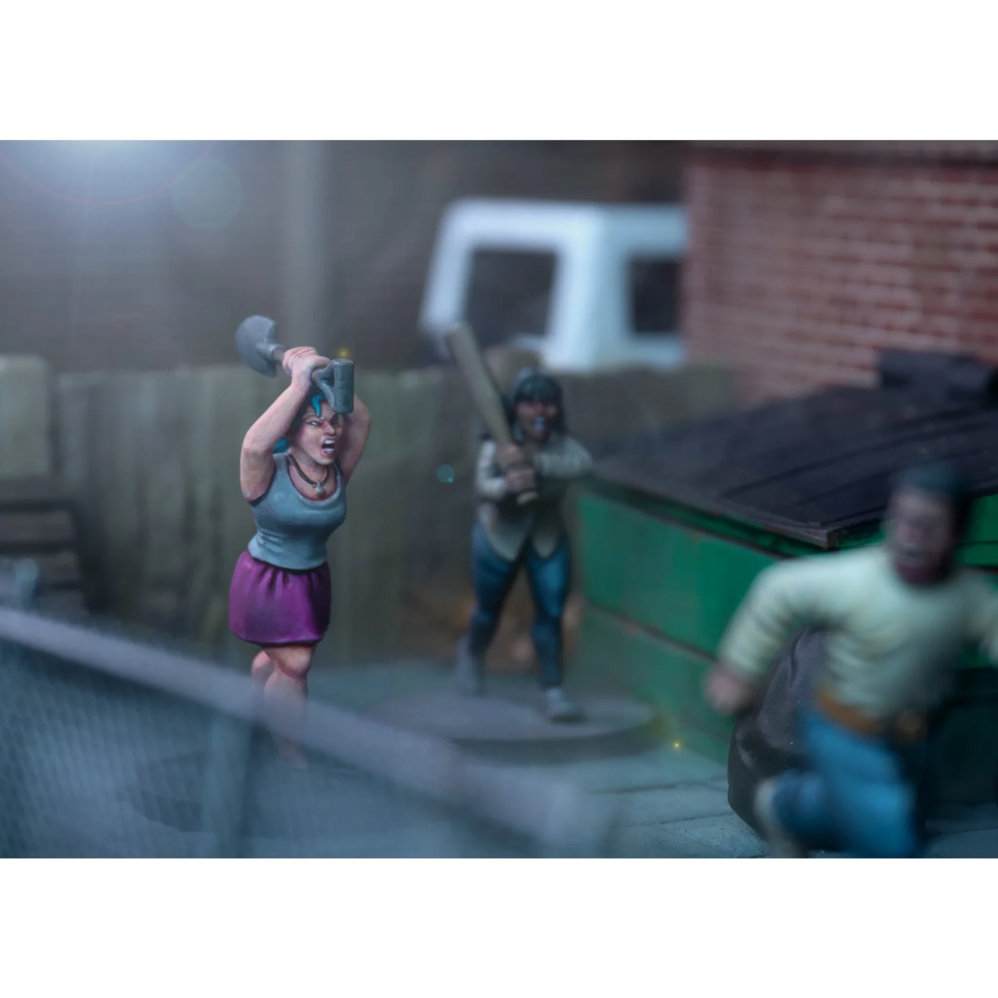 A dynamic scene features three miniature figures in a tense urban setting. In the foreground, a woman with blue hair raises a shovel, while a second figure behind her holds a baseball bat, both appearing ready to confront a threat. A third figure, dressed in a light shirt and blue pants, is seen running away, adding to the sense of urgency in the scene, with a green dumpster and a wooden fence in the background.
