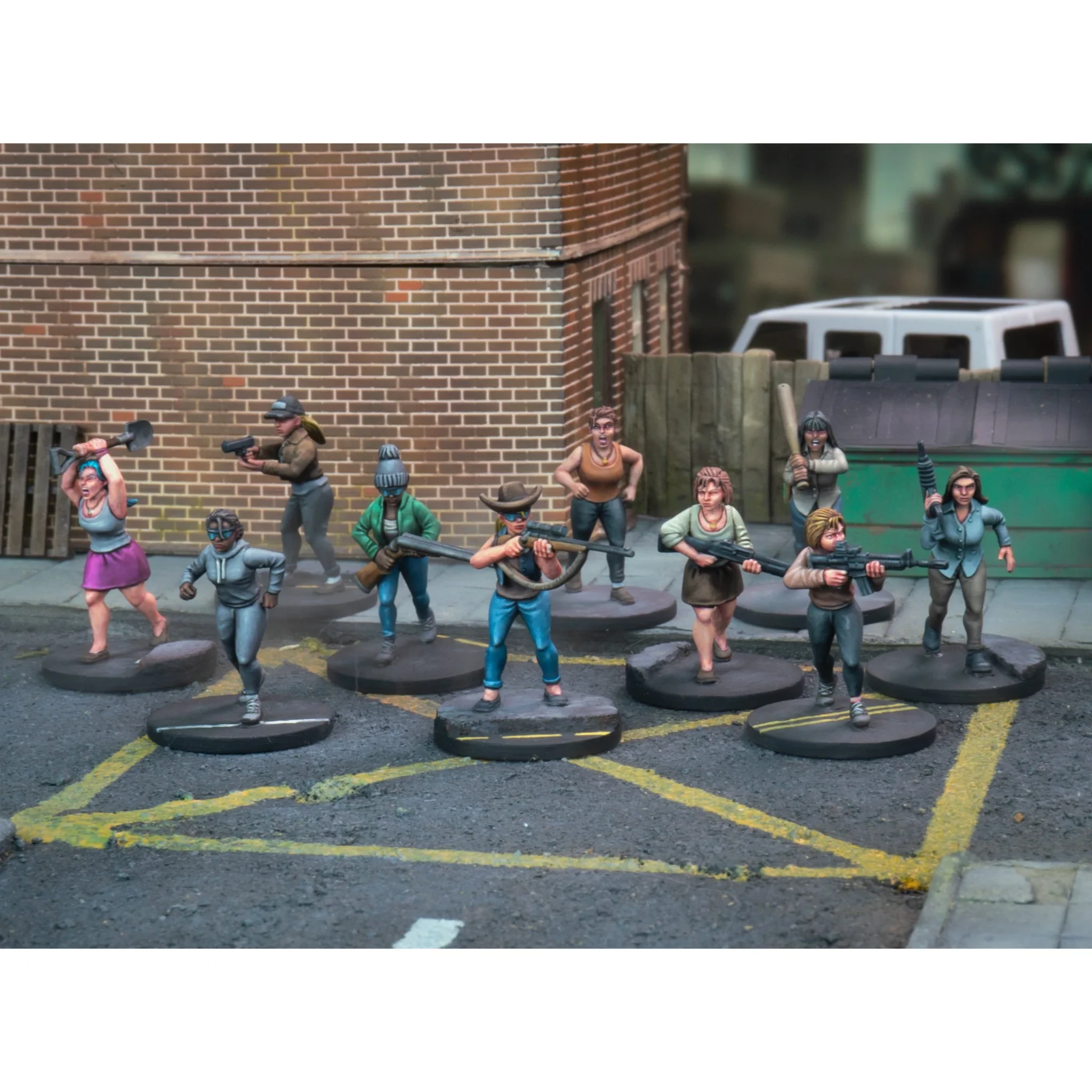 A group of ten miniature figures representing civilians and survivors stands on a street marked with yellow lines, set against a backdrop of a brick building and a dumpster. The figures display a variety of poses and expressions, holding weapons such as shovels, rifles, and baseball bats, showcasing a mix of panic and determination. The scene captures the essence of asymmetric conflict, with characters ranging from evacuees to hardened survivors, all intricately detailed and ready for tabletop gaming.