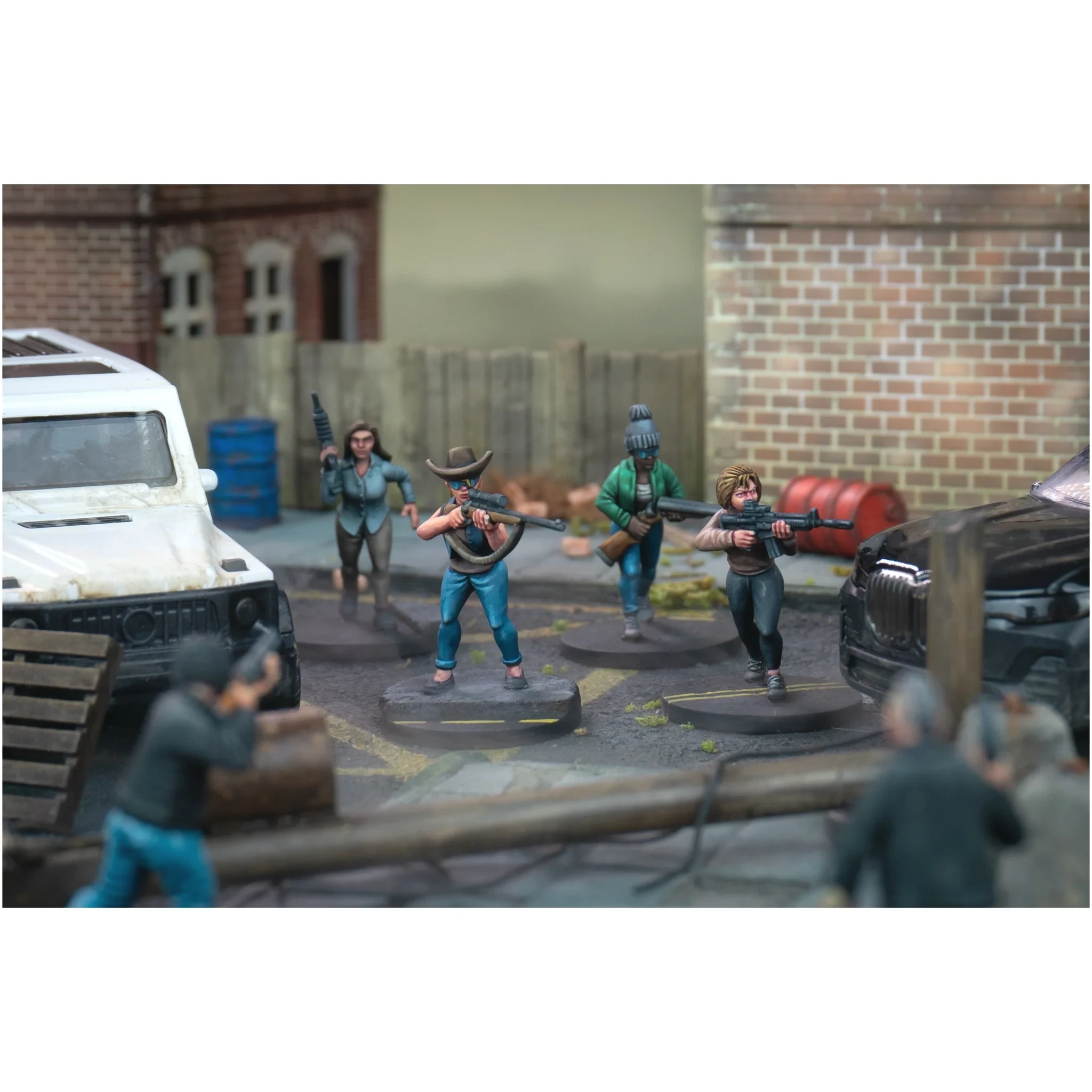 A dynamic scene features several miniature figures representing civilians and survivors in a post-apocalyptic setting. The figures are posed in various stances, some holding weapons like shotguns and rifles, while others appear to be in motion, suggesting a tense atmosphere. The backdrop includes a dilapidated urban environment with a white vehicle, barrels, and a wooden barricade, enhancing the sense of conflict and urgency.