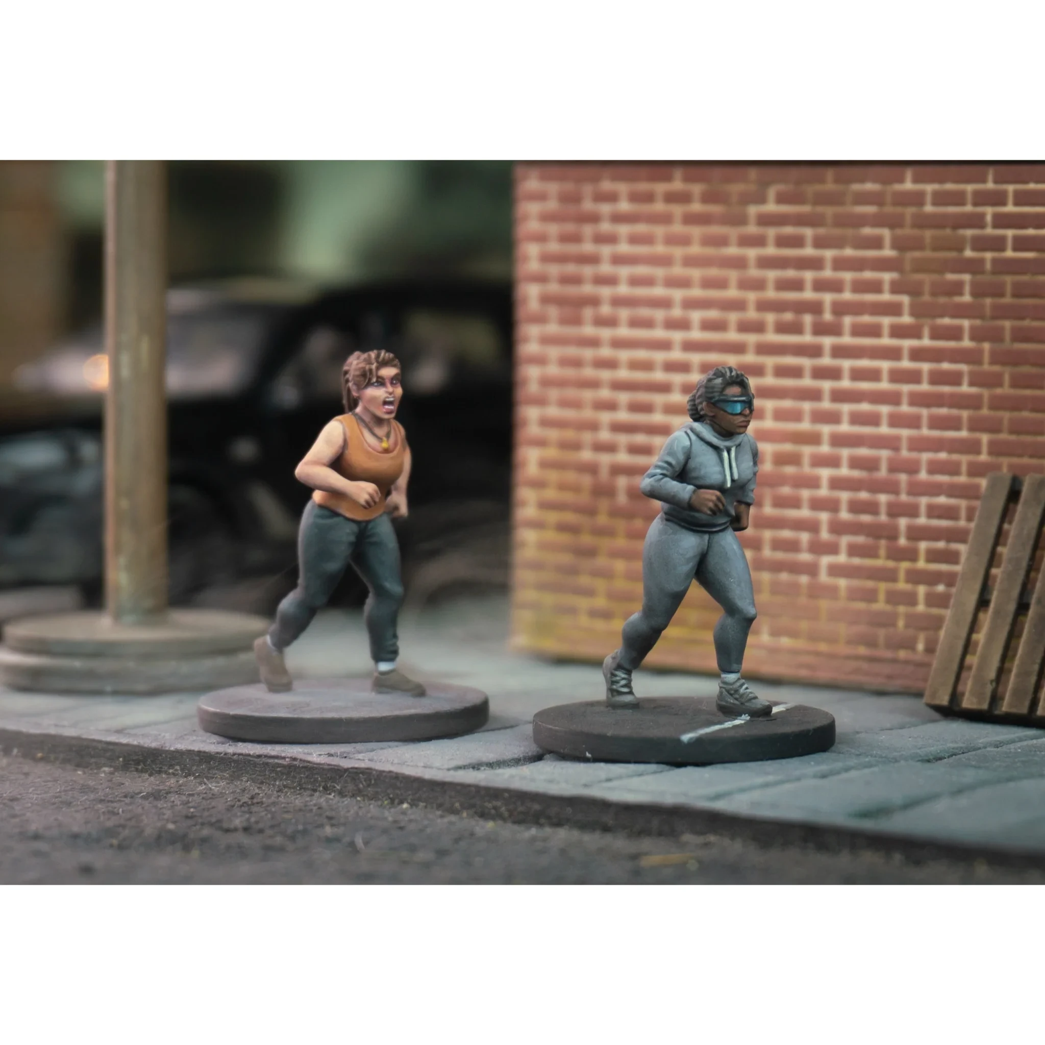 Two miniature figures are depicted on a cobblestone street, showcasing a scene of urgency. On the left, a woman with curly hair, wearing a brown tank top and dark pants, appears to be running with a startled expression. On the right, another woman in a gray tracksuit and a face mask is also in motion, suggesting a hurried escape. The background features a brick wall and a hint of urban elements, enhancing the atmosphere of a chaotic environment.