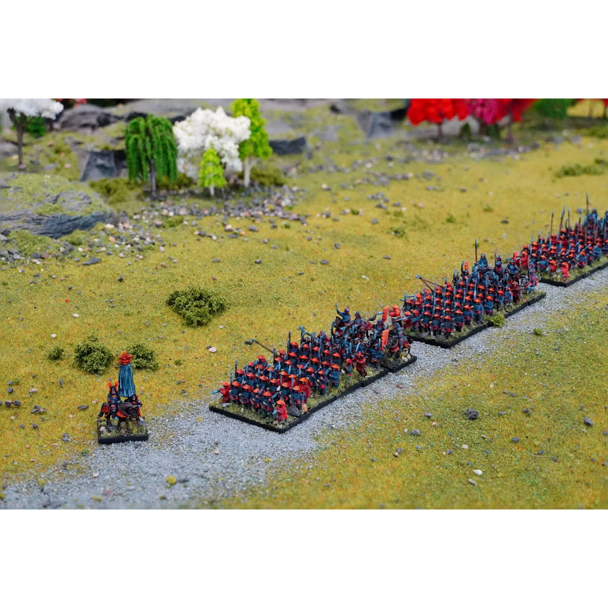 A detailed tabletop scene features a formation of 10mm miniatures representing Sengoku-era Japanese warriors. In the foreground, a single mounted figure in a blue cloak commands a line of Ashigaru soldiers, dressed in red and blue armor, standing in formation on a grassy battlefield. The background includes lush greenery and trees, enhancing the historical setting of the miniature display.