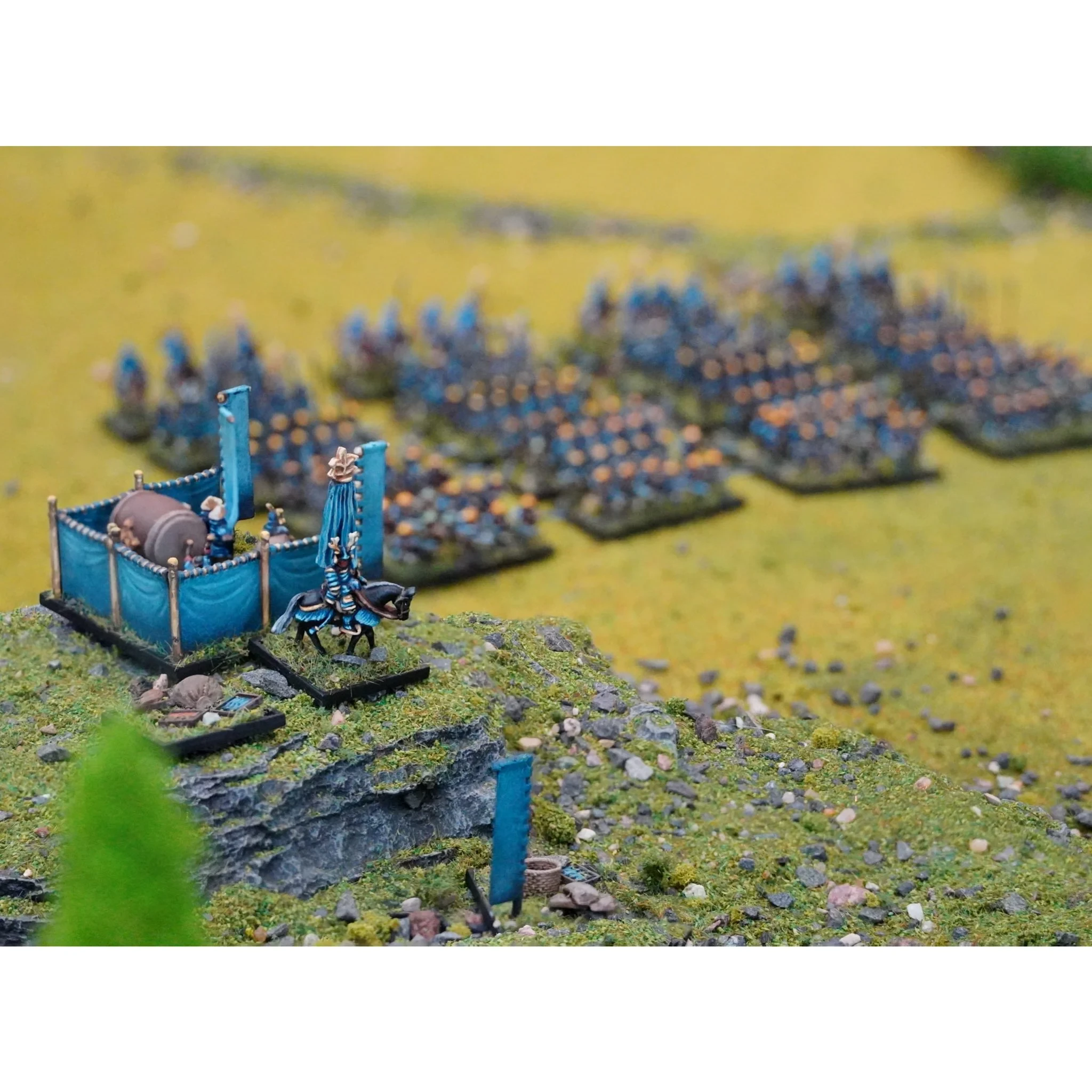 A detailed tabletop scene features a variety of 10mm miniatures representing Sengoku-era Japan. In the foreground, a small command unit with a horse and blue banners is positioned on rocky terrain, while in the background, numerous infantry figures in blue uniforms are arranged in organized formations on a grassy battlefield. The overall setting is vibrant, showcasing the intricate details of the miniatures and the lush green landscape.