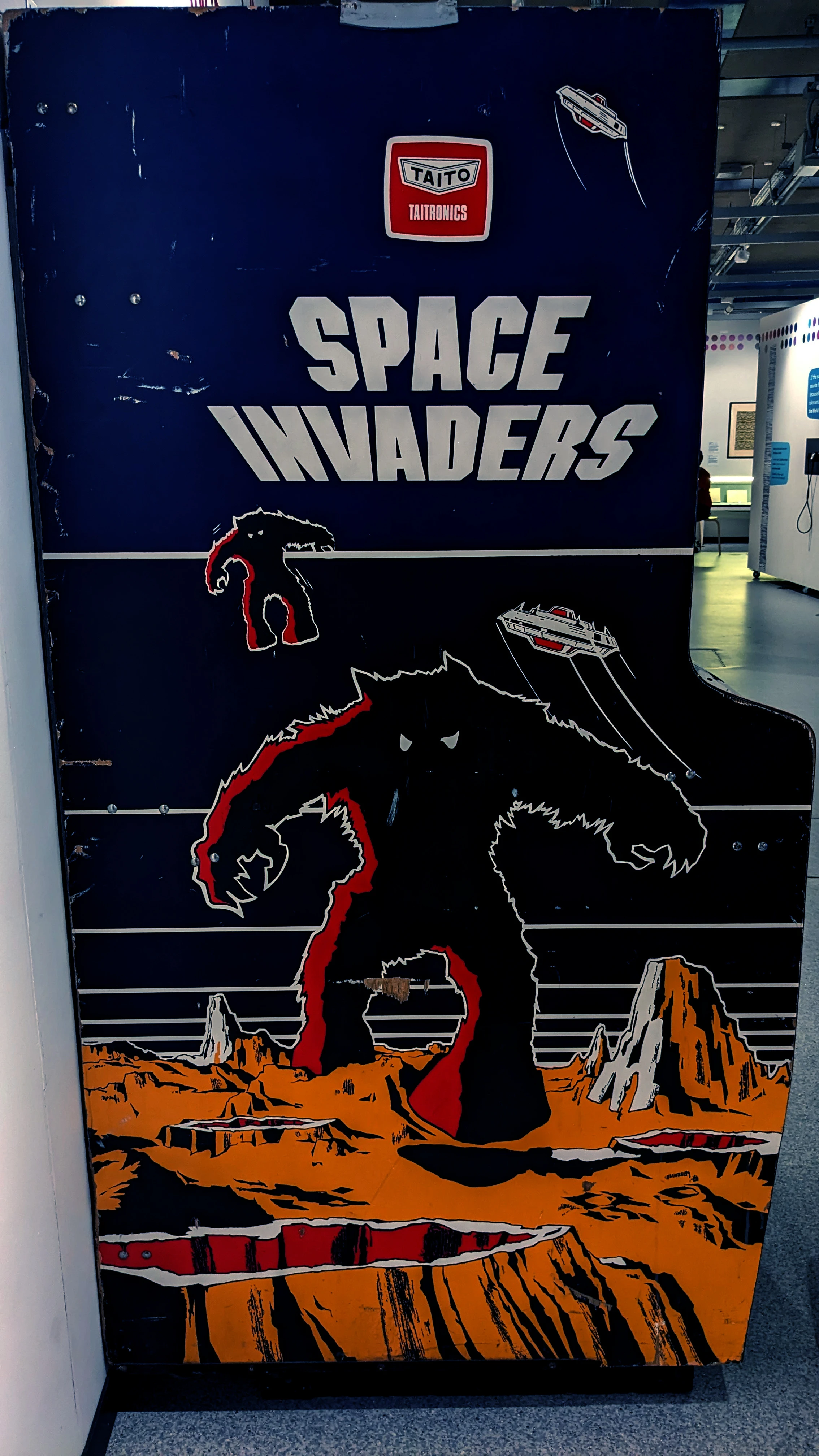 A vintage arcade cabinet for the game "Space Invaders" features a striking design with bold, white lettering at the top. The artwork depicts a large, menacing alien creature in black and red, with smaller figures and a spaceship above it, set against a dark blue background. The cabinet shows signs of wear, adding to its nostalgic charm, and is positioned in a well-lit space that suggests a gaming or exhibition environment.