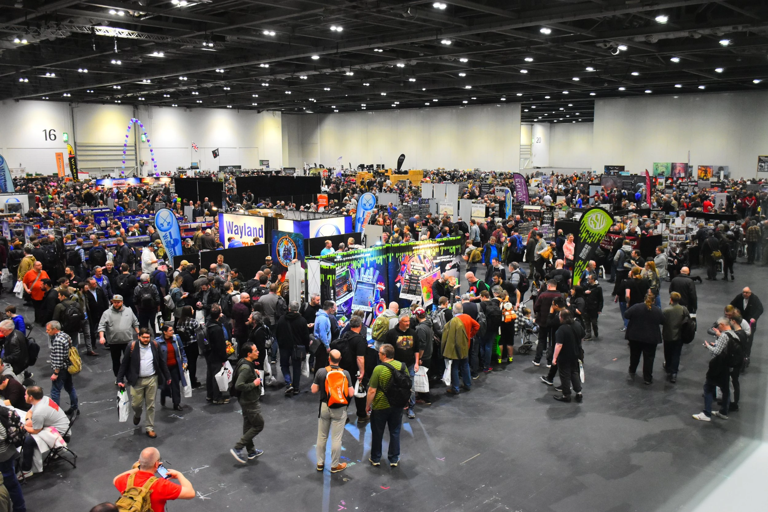 A bustling convention scene filled with a large crowd of attendees, many of whom are carrying bags. Various vendor booths are visible, including one for "Wayland Games," with colorful banners and displays. The space is well-lit, showcasing a mix of people in casual attire, some wearing backpacks, and a variety of promotional signage in the background.