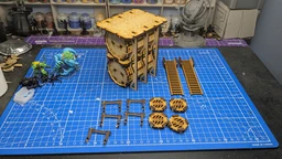 A detailed assembly of the TT Combat Sector 4 Storage tanks is displayed on a blue cutting mat marked with a grid. The main structure consists of two large, cylindrical tanks stacked vertically, supported by a wooden frame with intricate detailing. Surrounding the tanks are various components, including two sets of ladders, several hazard markers labeled "DANGER," and additional structural pieces, all crafted from a light wood material. In the background, a miniature figure and a globe stand are visible, adding context to the tabletop gaming environment.