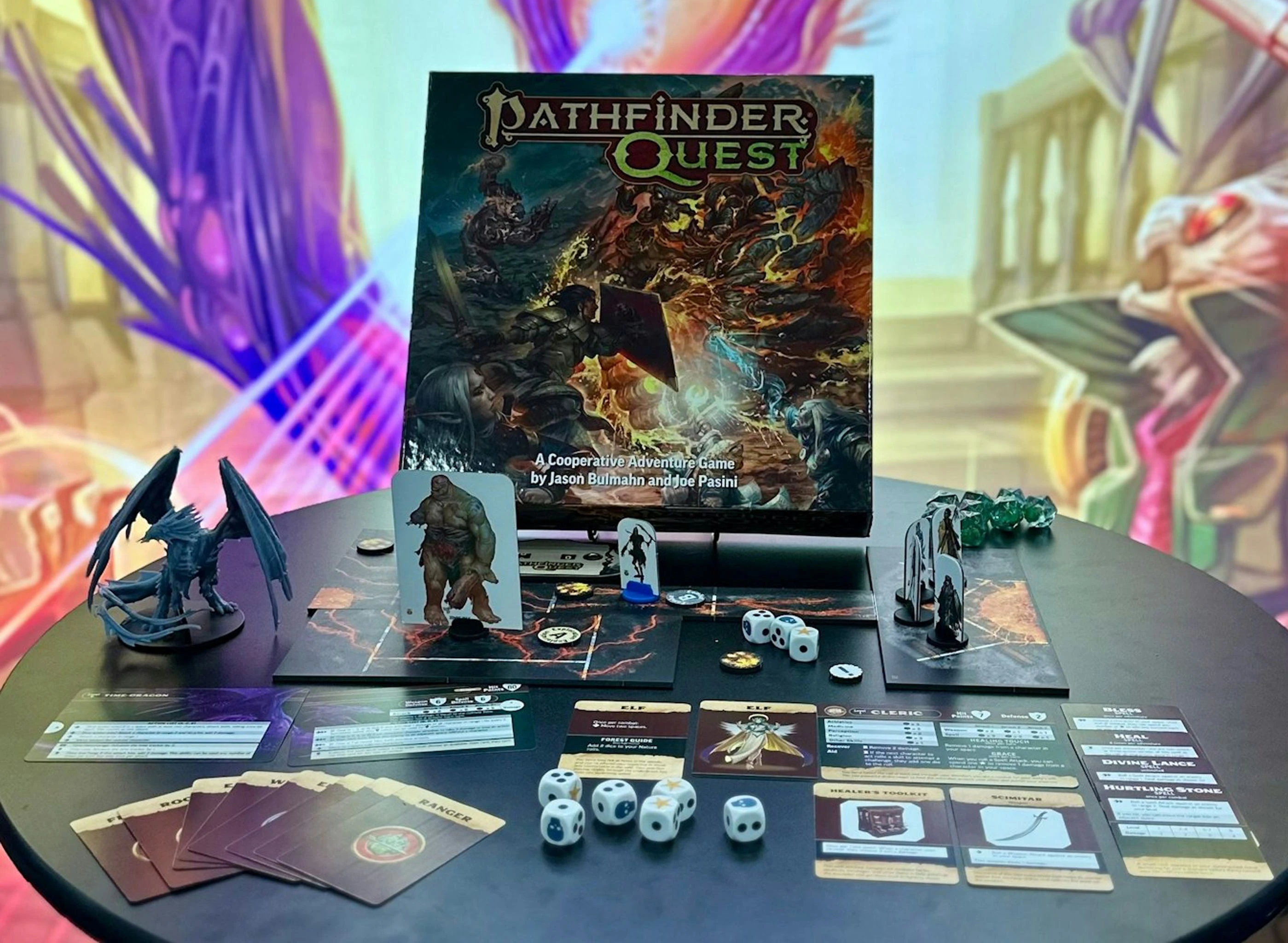 A tabletop display features the "Pathfinder Quest" game box prominently in the center, showcasing dynamic artwork of characters in battle. Surrounding the box are various game components, including character standees, dice, and cards representing different classes like Ranger and Cleric. The scene is set on a dark surface, with a colorful, abstract background adding depth to the presentation.
