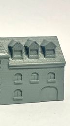 A detailed model of a building designed for 6mm scale wargames, featuring a sloped roof with three dormer windows and a textured surface that resembles slate. The facade includes several arched and rectangular windows, along with a large door at the base, all rendered in a light gray resin. The overall design suggests an industrial style, suitable for various historical or modern warfare settings.