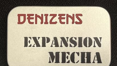 A small, silver tin features the text "DENIZENS" in bold red letters at the top and "EXPANSION MECHA" in large, black, stencil-style font below. The tin's surface has a smooth, metallic finish, and it is set against a dark background, emphasizing its compact and portable design. This packaging represents the new expansion for the Space Denizens wargame, which includes mechs and terrain features.
