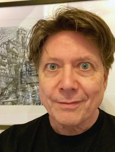 A close-up portrait of a man with light brown hair and striking green eyes, smiling slightly. He is wearing a black shirt and is positioned in front of a detailed, artistic background featuring a complex, fantastical structure, possibly a building or landscape. The overall tone of the image is casual and inviting, with a focus on the subject's expressive features.