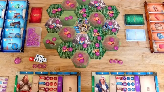 Board game setup with hexagonal green terrain tiles featuring pink and purple structures. Miniature figures and small tokens scattered across. Two player boards with character portraits, resource dials, and action icons. Dice and small bags of colored tokens on the table.
