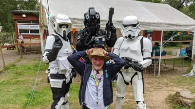A man in a wide-brimmed hat and a casual outfit kneels in the foreground, smiling and playfully raising his hands above his head. Behind him, two Stormtroopers and a TIE Fighter pilot in full costume stand with their weapons, creating a fun and engaging atmosphere. The setting features a grassy area with a tent in the background, suggesting a lively outdoor event.