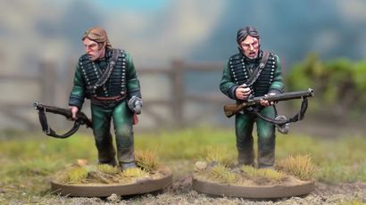 Two detailed miniatures of soldiers from the Sharpe’s Rifles series are displayed on textured bases, set against a blurred natural background. Both figures wear green uniforms with black crossbelts; one holds a rifle and has a serious expression, while the other appears to be in a more contemplative pose. The bases feature tufts of grass, enhancing the realistic terrain effect.