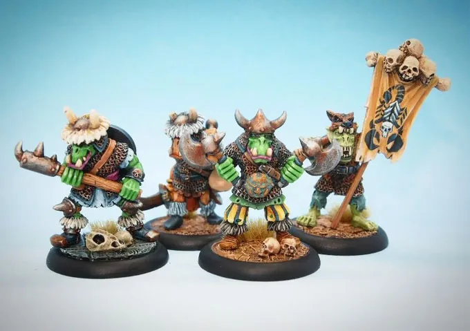 Four intricately painted orc miniatures stand against a light blue background. The first orc on the left wields a spiked mace and has a skull at its feet, while the second orc, wearing a horned helmet, holds a large axe. The third orc, prominently displaying green skin and wearing a decorated tunic, gestures with its hands, and the fourth orc carries a banner adorned with skulls and a scorpion motif. Each figure showcases detailed armor and accessories, highlighting their unique characteristics.