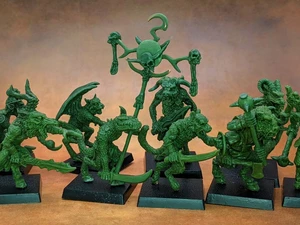 A collection of old school miniatures from Satyr Art Studio, featuring various fantastical creatures in a vibrant green color. The figures include beastmen with distinct features such as horns and claws, some wielding weapons, and a central figure holding a staff topped with a skull and a crescent. They are displayed on black bases against a warm, textured background.