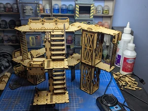A detailed assembly of modular terrain pieces made from laser-cut wood, featuring multiple levels, staircases, and platforms. The structure includes various textures and patterns, with a mix of open and enclosed areas, showcasing a futuristic industrial design. In the background, paint bottles and tools are visible, indicating a workspace for painting and customizing the terrain.