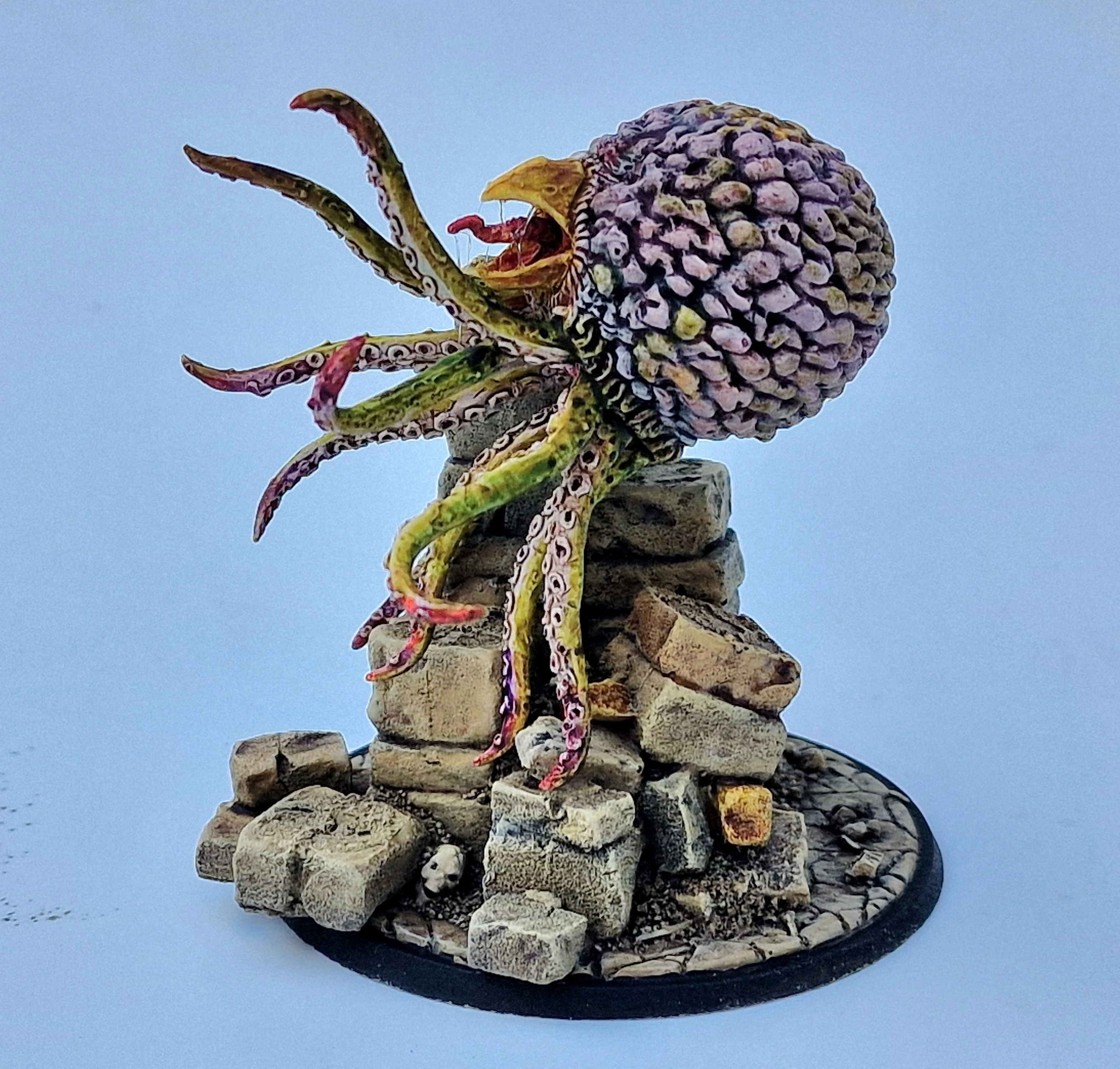 A detailed miniature of a creature resembling a large, bulbous head with a textured surface, featuring vibrant colors like purple and green. It has multiple long, tentacle-like appendages extending from its base, which is perched atop a pile of irregularly shaped stone blocks. The base is circular and textured, resembling a rocky terrain, enhancing the overall fantasy aesthetic of the piece.