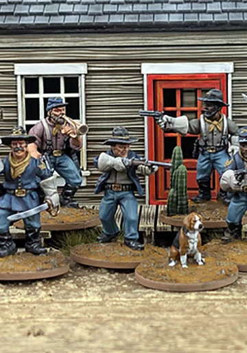 A group of ten detailed 28mm scale miniatures representing dismounted US Cavalry and Buffalo Soldiers stands in front of a wooden building with a red door. The miniatures are posed with various weapons, including rifles and a bugle, showcasing a mix of uniforms and accessories, such as scarves and hats. A small beagle and a cactus are also present among the figures, all mounted on 25mm round bases with a sandy terrain effect.