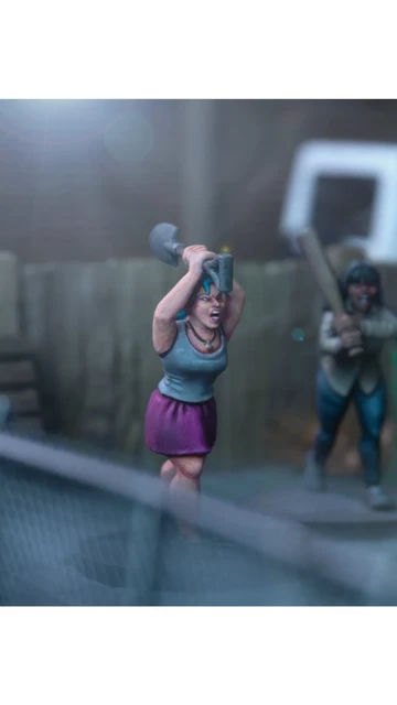 A dynamic scene features three miniature figures in a tense urban setting. In the foreground, a woman with blue hair raises a shovel, while a second figure behind her holds a baseball bat, both appearing ready to confront a threat. A third figure, dressed in a light shirt and blue pants, is seen running away, adding to the sense of urgency in the scene, with a green dumpster and a wooden fence in the background.