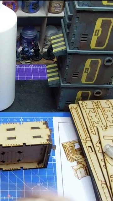 A workbench scene features a partially assembled laser-cut building structure made of light wood, positioned in the foreground. Behind it, a stack of completed structures in various colors, including blue and yellow, is visible, alongside bottles of paint and glue. A cutting tool rests on the table, and a printed reference image of a similar structure is partially visible.