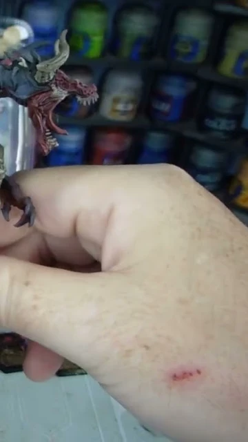 A detailed dragon miniature is being held in a hand, showcasing its intricate features, including prominent scales, fierce facial expression, and large wings with a red underside. The dragon is painted in dark hues with highlights that accentuate its muscular form and menacing appearance. In the background, various paint jars are visible, indicating a workspace for model painting.