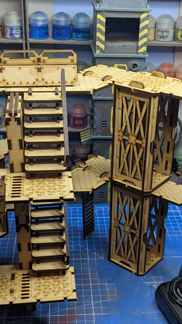A detailed assembly of modular terrain pieces made from laser-cut wood, featuring multiple levels, staircases, and platforms. The structure includes various textures and patterns, with a mix of open and enclosed areas, showcasing a futuristic industrial design. In the background, paint bottles and tools are visible, indicating a workspace for painting and customizing the terrain.
