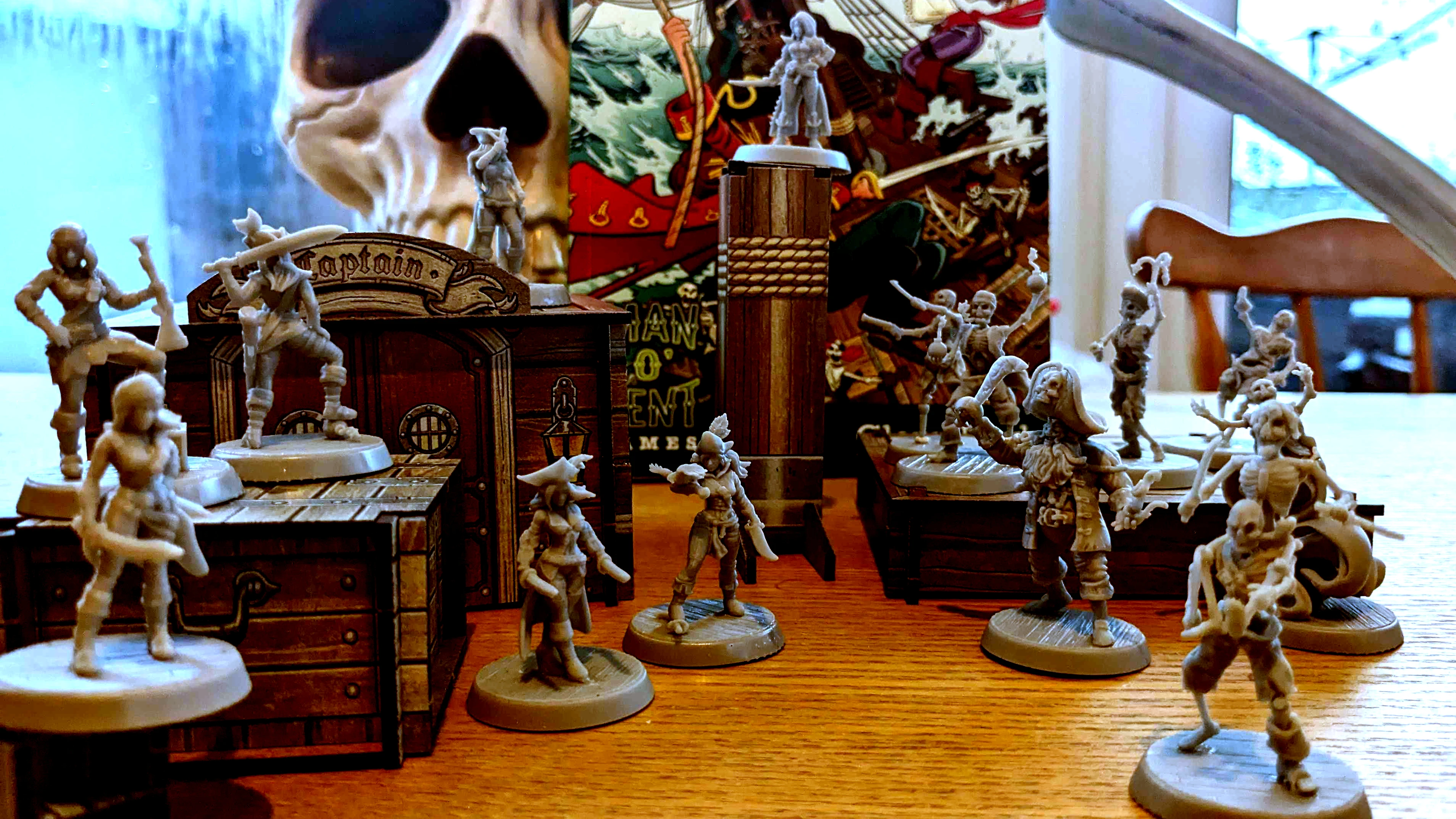 A collection of detailed miniatures from the game "Boarders and Black Flags" is displayed on a wooden surface, featuring both human pirates and undead Skelliwags. The miniatures are unpainted and vary in poses, with some wielding swords and others appearing to engage in action. In the background, a vibrant pirate-themed backdrop showcases a skull and crossbones design, along with elements of a ship, enhancing the thematic atmosphere of the game.