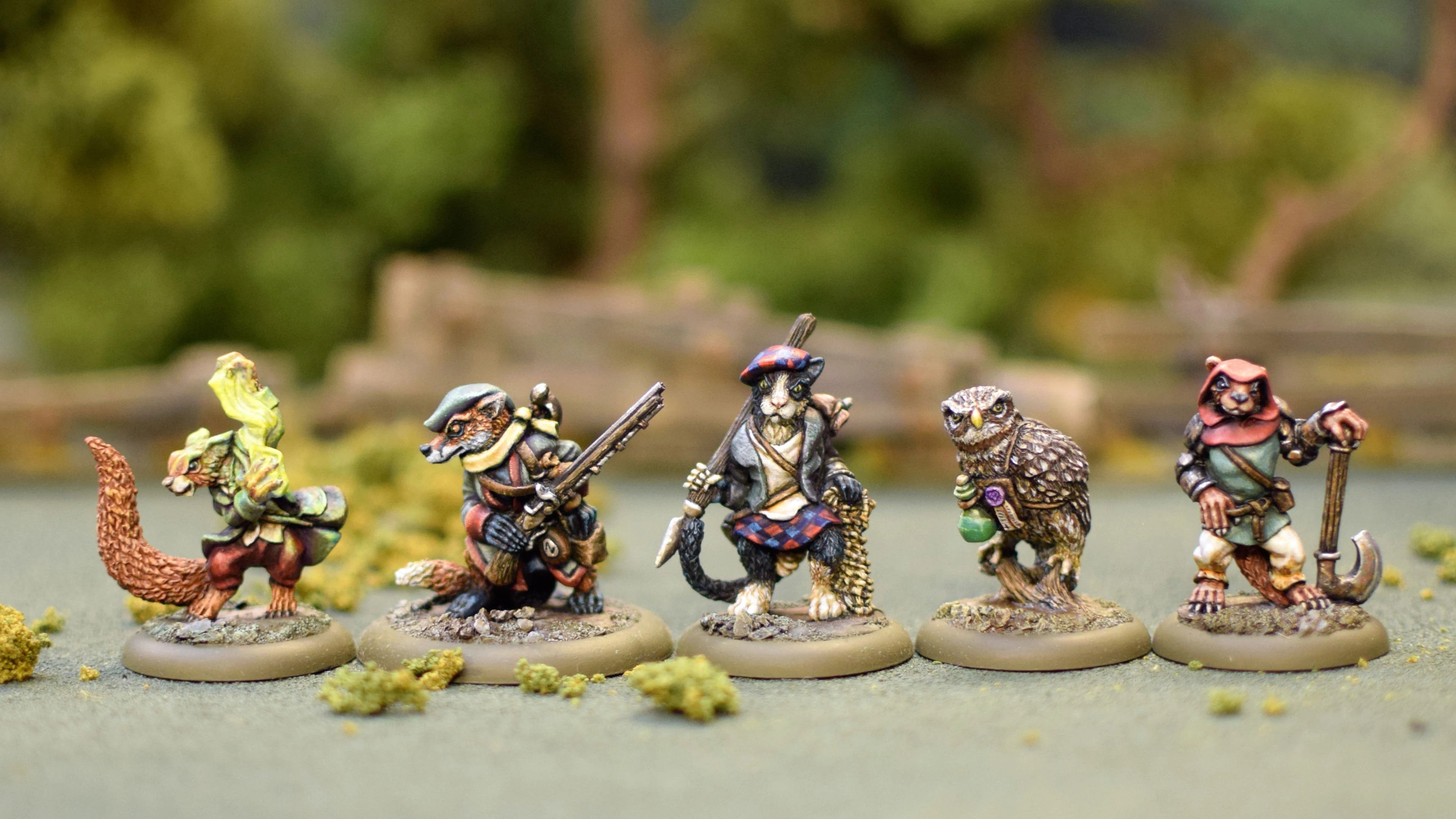 A group of four anthropomorphic miniatures from the game Burrows and Badgers, each character displaying unique features and attire. On the left, a badger holds a rifle, dressed in a green and brown outfit. Next to it, a cat in a blue and red tartan outfit wields a sword. An owl stands in the center, holding a potion, while a rabbit on the right wears a red cloak and holds a dagger. The miniatures are set against a blurred natural background, enhancing their detailed sculpting and painting.