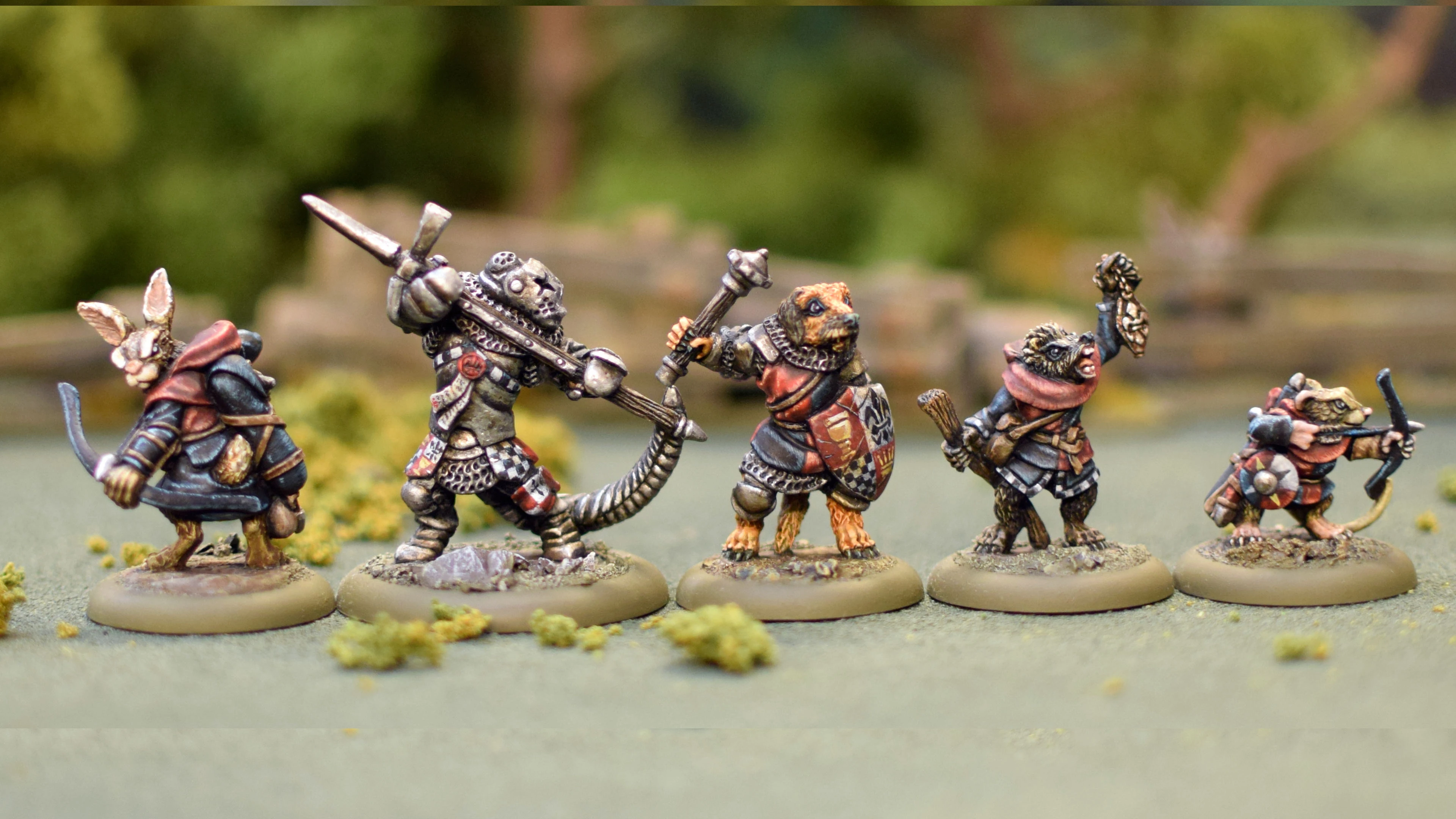 Four anthropomorphic animal miniatures are depicted in a dynamic scene, showcasing characters from the game Burrows and Badgers. The central figure, a fierce-looking warrior with a spiked weapon, is flanked by a bear in armor and two other characters, one holding a staff and the other wielding a sword. The miniatures are intricately detailed, with a variety of colors and textures, set against a blurred natural background that suggests a battlefield or forest setting.