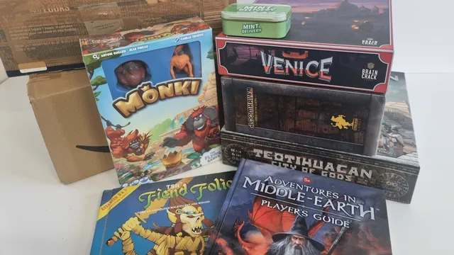 Stack of board games and books, including "MØNKI", "VENICE", "Adventures in Middle-Earth", and "Fiend Folio".