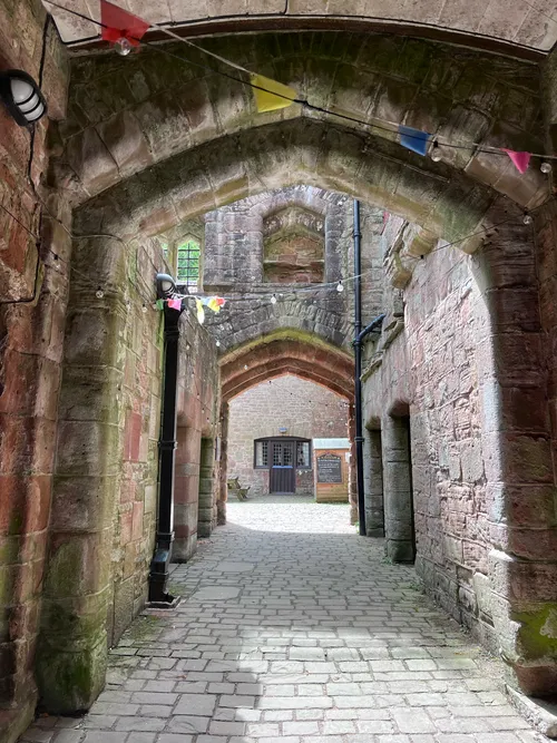 A stone archway leads into a narrow passageway within a medieval castle, featuring weathered red stone walls and a cobblestone floor. Colorful triangular flags hang above, adding a festive touch, while soft lighting from wall-mounted fixtures illuminates the scene. In the distance, an open doorway reveals a glimpse of the castle's interior, enhancing the atmospheric and slightly haunted vibe of the setting.