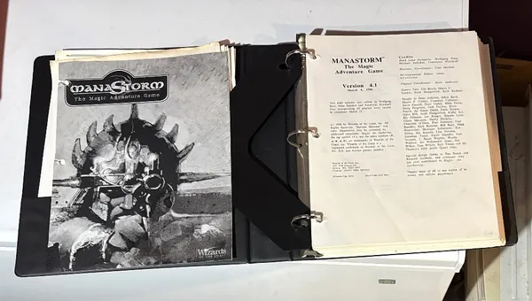 A three-ring binder containing the playtest manuscript for "ManaStorm: The Magic Adventure Game," featuring a black and white cover illustration of a character with a spiked helmet and a weapon. The title "MANASTORM The Magic Adventure Game" is prominently displayed at the top, along with the version number and date on the first page inside the binder. Additional pages include loose character sheets and player handouts, indicating the game's mechanics and spells.