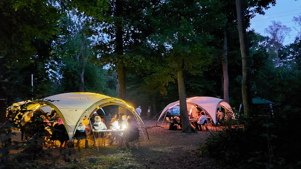 A cozy outdoor gaming setup features several large, white tents illuminated by warm lights, surrounded by trees in a wooded area during dusk. Groups of people are seated at tables under the tents, engaged in games and conversation, creating a relaxed and social atmosphere. The ground is covered in leaves, and the soft glow from the lights adds to the inviting ambiance of the gathering.