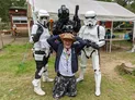 A man in a wide-brimmed hat and a casual outfit kneels in the foreground, smiling and playfully raising his hands above his head. Behind him, two Stormtroopers and a TIE Fighter pilot in full costume stand with their weapons, creating a fun and engaging atmosphere. The setting features a grassy area with a tent in the background, suggesting a lively outdoor event.