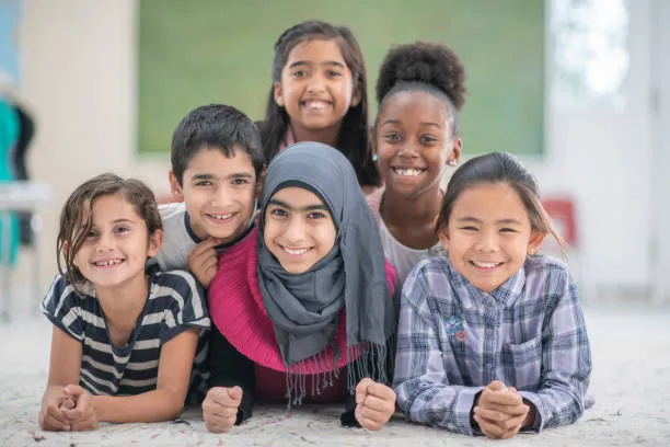 A group of six children is smiling and posing together on a light-colored carpet. They are arranged in a semi-circle, with the girl in the front wearing a pink sweater and a grey hijab, while the others are dressed in various colorful outfits. The background features a softly blurred interior space, suggesting a warm and welcoming environment.