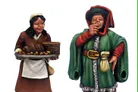 A miniature figure of a woman in a brown dress and white apron holds a tray filled with loaves of bread, smiling as she stands on the left. Beside her, a man in a colorful green cloak and red hat raises a cup to his lips, wearing a sly expression. Both figures are intricately detailed, showcasing the vibrant colors and textures typical of fantasy tavern patrons.