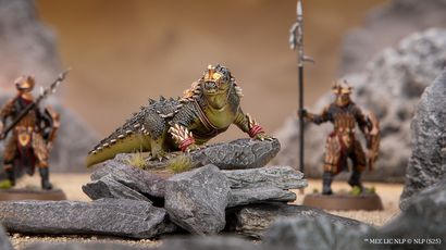 A fierce, green war drake with prominent spines and decorative armor stands atop a rocky outcrop, showcasing its detailed scales and menacing expression. In the background, an Easterling warrior clad in ornate armor holds a spear, partially obscured by rocks, emphasizing the drake's imposing presence. The scene is set against a blurred, sandy terrain, enhancing the focus on the miniatures.