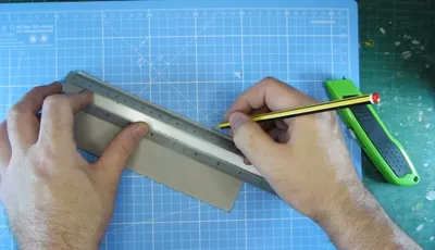 A hand is holding a yellow pencil against a metal ruler, positioned on a piece of cardboard on a cutting mat. The cutting mat features a grid pattern with measurements in centimeters, and a green utility knife is visible nearby. The scene suggests preparation for cutting or crafting a new paint shelf.