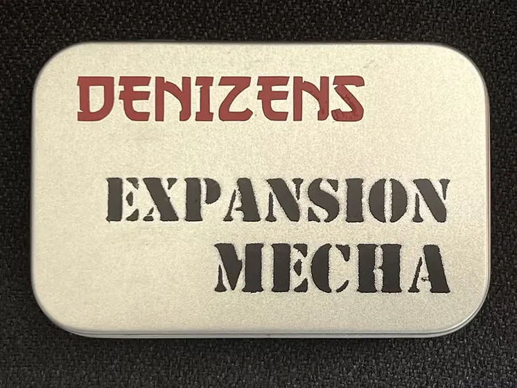 A small, silver tin features the text "DENIZENS" in bold red letters at the top and "EXPANSION MECHA" in large, black, stencil-style font below. The tin's surface has a smooth, metallic finish, and it is set against a dark background, emphasizing its compact and portable design. This packaging represents the new expansion for the Space Denizens wargame, which includes mechs and terrain features.