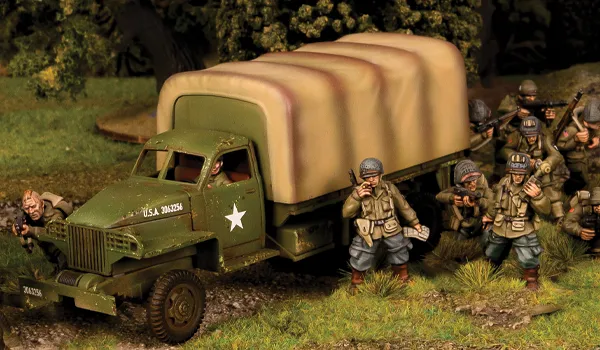 A military scene features a green WWII truck with a large, canvas-covered cargo area, displaying a white star insignia on its side. Surrounding the truck are several soldiers in period-appropriate uniforms, some aiming their weapons and others in various poses, set against a grassy terrain with trees in the background. The overall atmosphere conveys a sense of readiness and action typical of wartime scenarios.